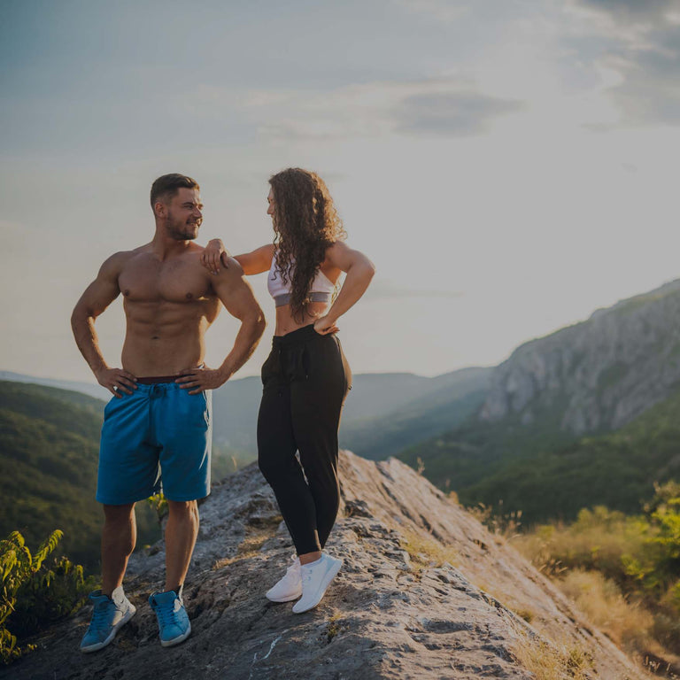 Two people in athletic wear standing on a rocky mountain ridge with a scenic valley and hills in the background during golden hour.