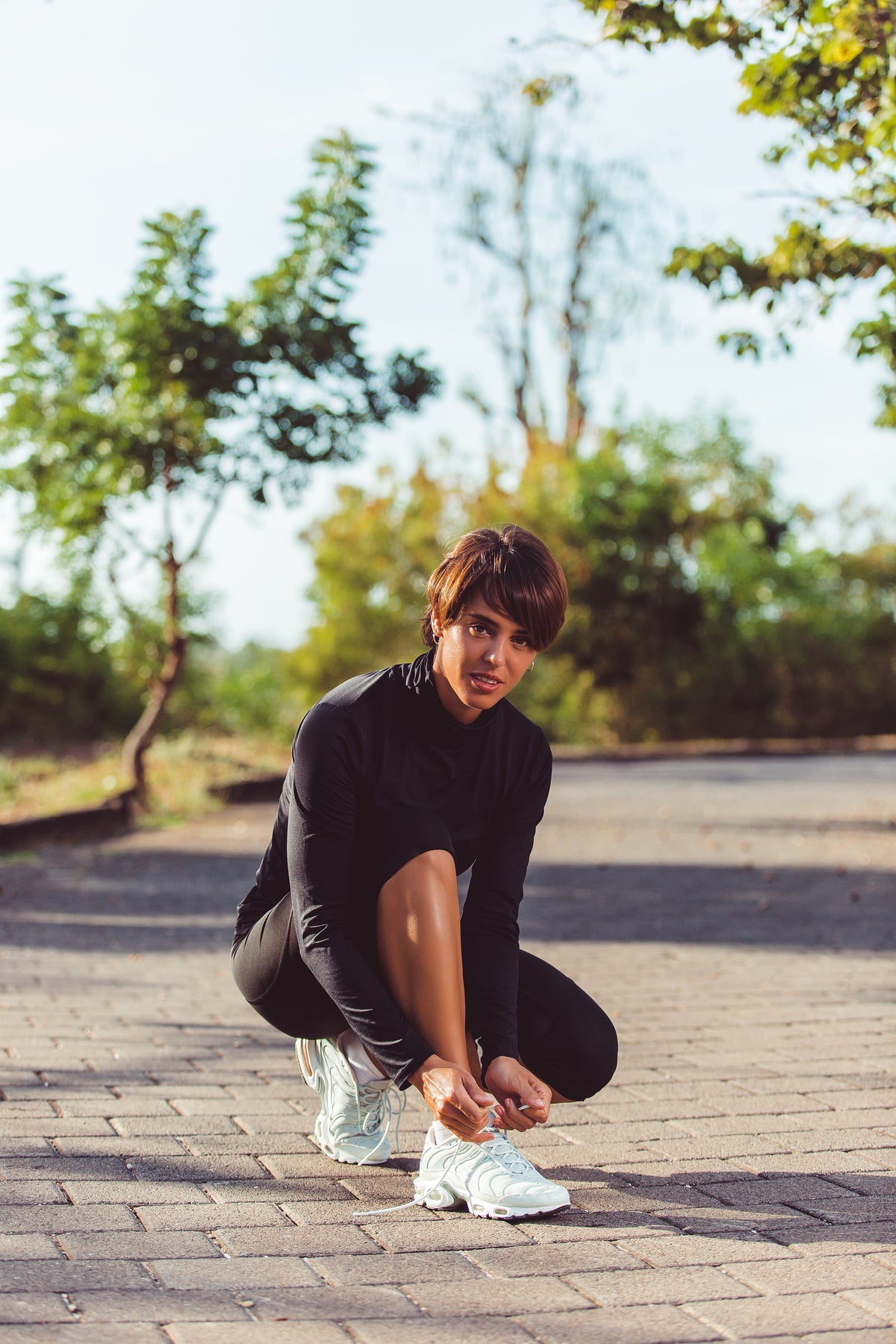 A woman in a black athletic long-sleeve top and black leggings crouches on a paved outdoor path, tying the laces of her white running shoes. Trees and greenery are blurred in the background.