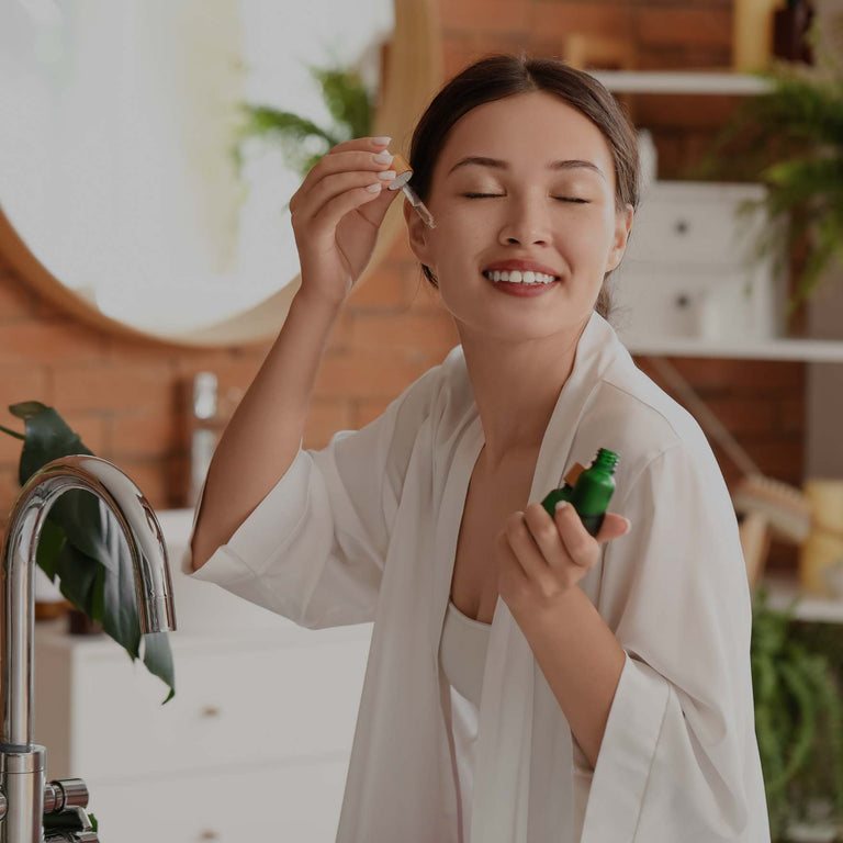 A woman in a white robe stands in a bathroom, holding a small green bottle and applying a drop of serum from a dropper to her face. The scene is warmly lit with a brick wall, mirror, and potted plants in the background.