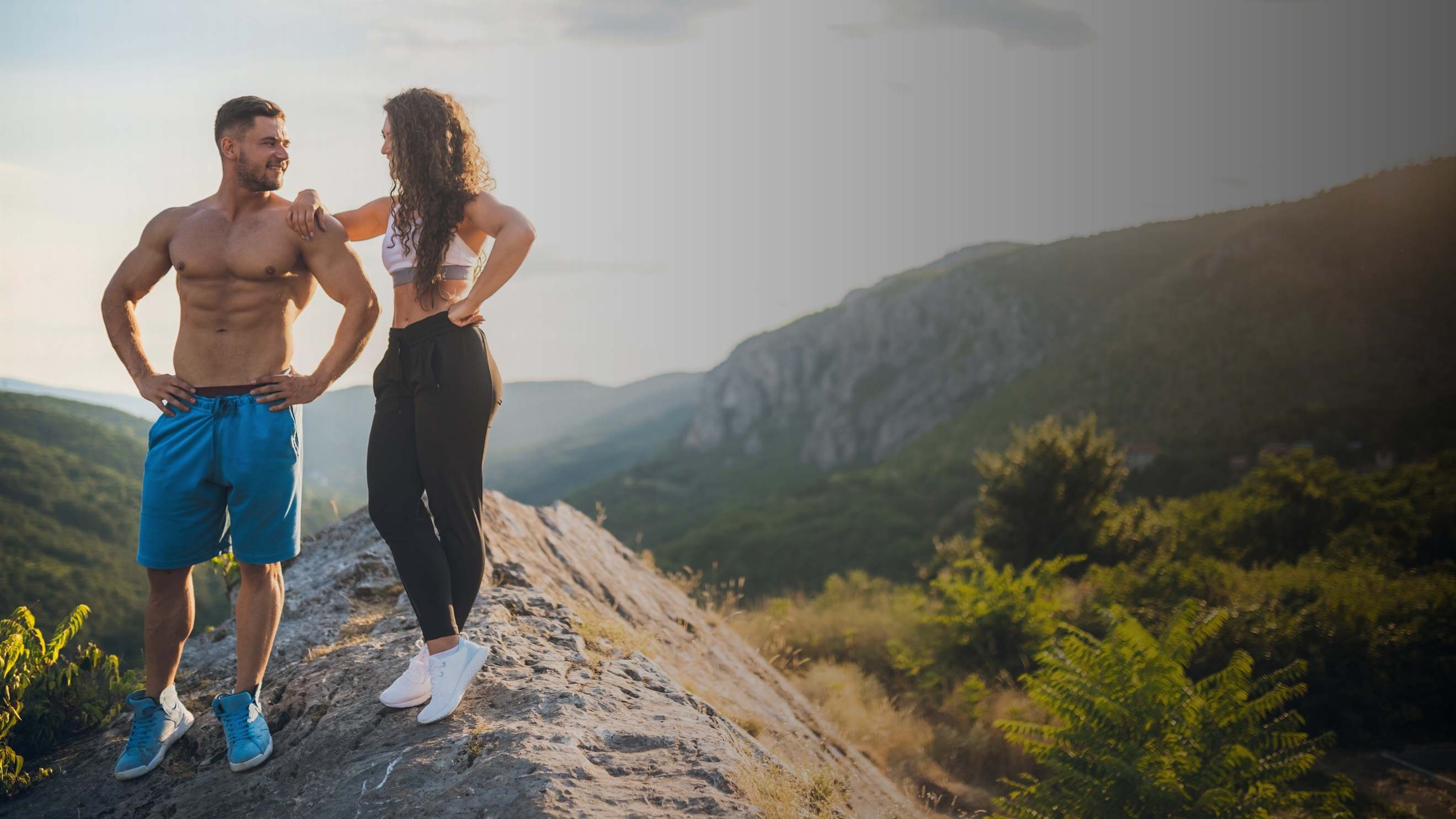 Two people in athletic wear standing on a rocky mountain ridge with a scenic valley and hills in the background during golden hour.
