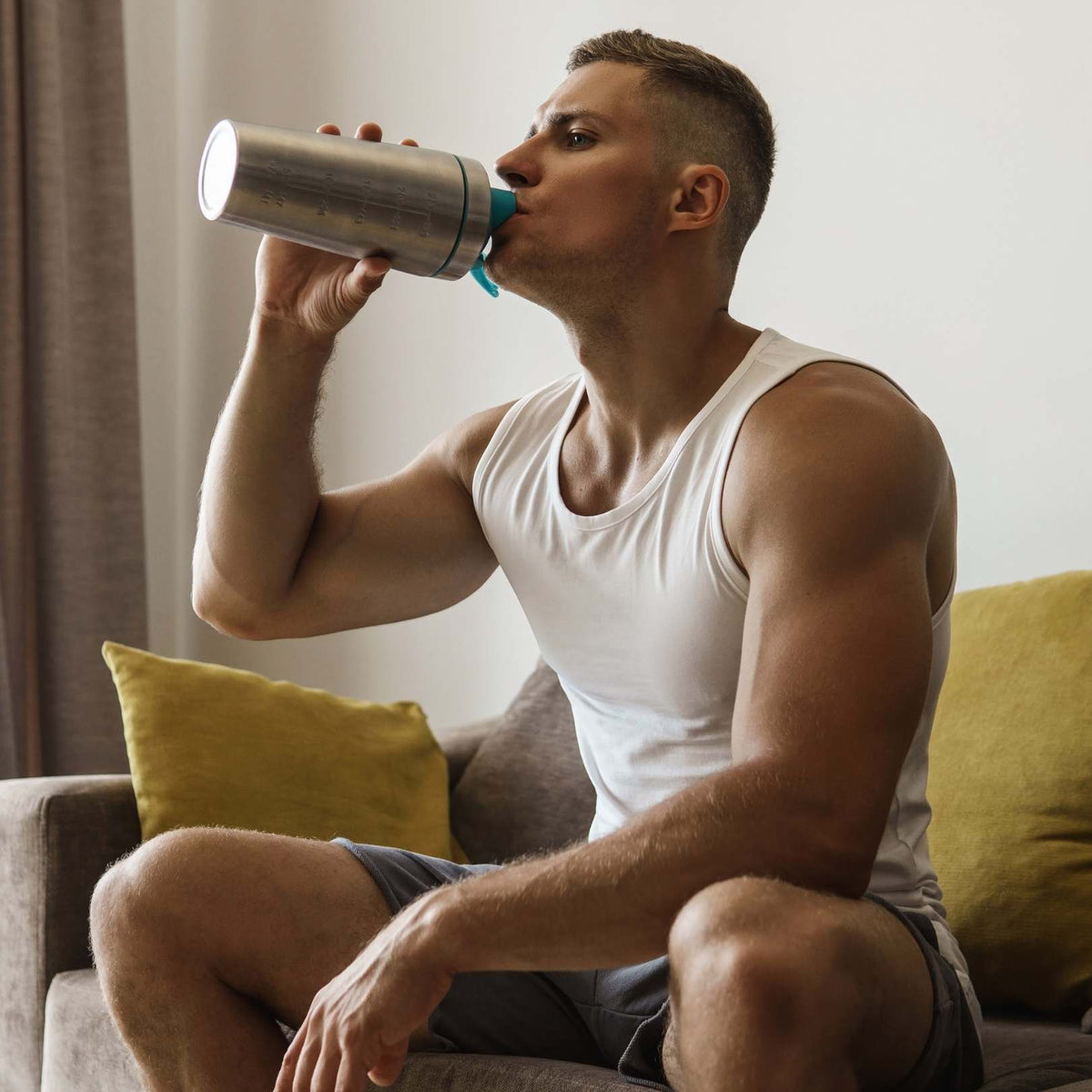 A man in a white tank top sitting on a couch with yellow pillows, drinking from a stainless steel water bottle with a blue cap.