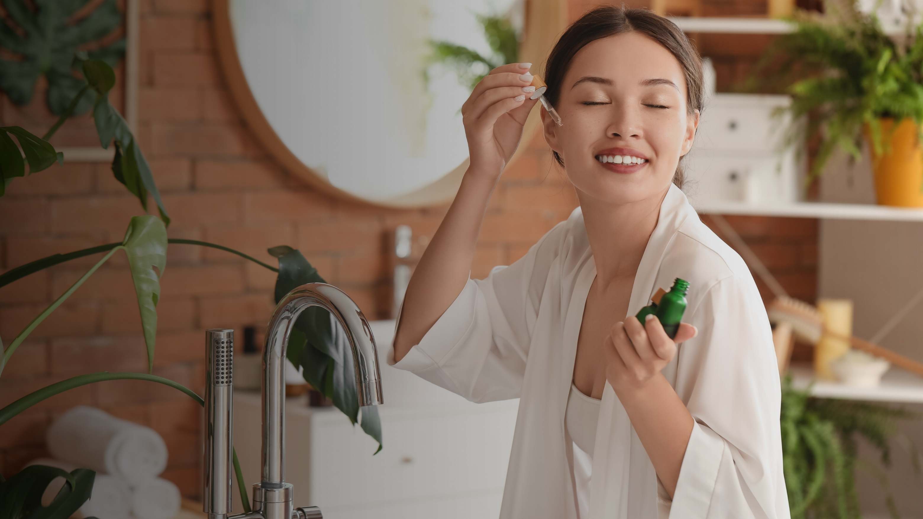 A woman in a white robe stands in a bathroom, holding a small green bottle and applying a drop of serum from a dropper to her face. The scene is warmly lit with a brick wall, mirror, and potted plants in the background.