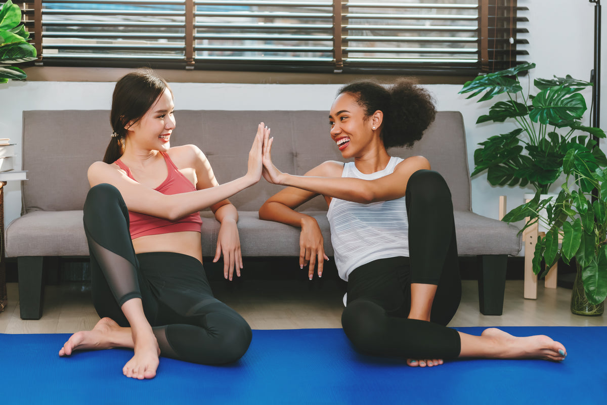 Two women in activewear sitting on a blue yoga mat in a living room, high-fiving in front of a gray sofa and green plants.